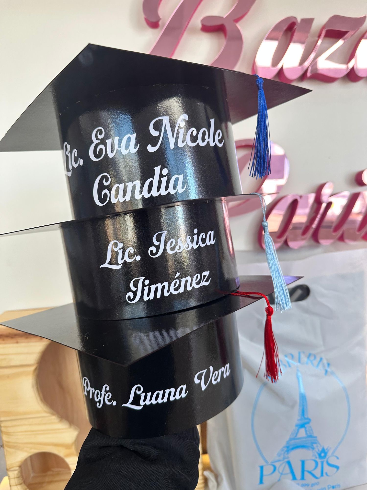 Birretes de graduación personalizados con nombres, apoyados frente al cartel de Bazar París